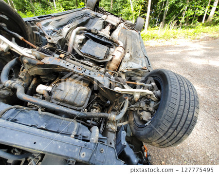 An overturned vehicle rests on its roof amidst trees gravel, indicating recent accident in quiet location. An overturned vehicle rests on its roof amidst trees gravel, indicating recent accident in quiet location. 127775495