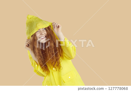 Happy young woman hiding from rain under yellow raincoat on beige copy space background 127778068