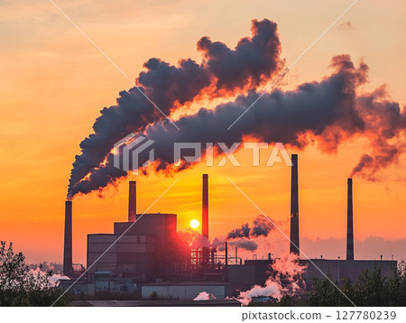 A factory with multiple smokestacks spews thick plumes of smoke against a fiery orange and red sunset sky, highlighting industrial pollution. 127780239