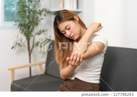 Elbow Pain Management. Young woman rubbing her elbow in discomfort at home. Elbow Pain Management. Young woman rubbing her elbow in discomfort at home. 127786263