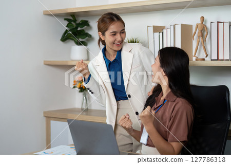 Celebrating Success in the Office. Two young women rejoice over project achievements in a modern workspace. 127786318
