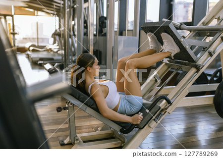 Beautiful woman sporty girl exercising using leg press machine at the gym Beautiful woman sporty girl exercising using leg press machine at the gym 127789269