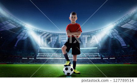 Young soccer player standing confidently on lit stadium field with ball. Youth sports ambition. 127789514