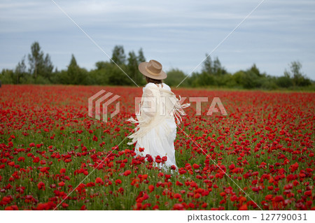 Young woman in a white dress and hat walks among summer poppies 127790031