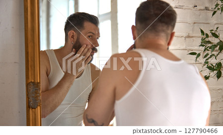 Clean-boy care. Man in casual loungewear applies gel to his face in front of the mirror. Clean-boy care. Man in casual loungewear applies gel to his face in front of the mirror. 127790466