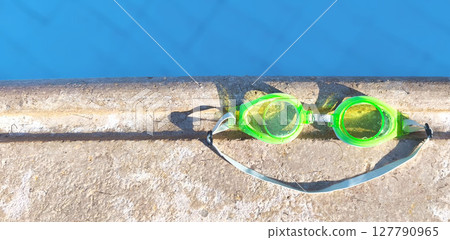 Bright green swimming goggles rest on the edge of a pool, casting a smile-shaped shadow in the sunlight. Perfect summer vibe and vacation mood 127790965