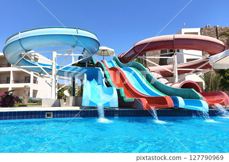Bright and colorful water slides splash into a crystal-clear blue pool on a sunny day at a resort, capturing the essence of summer fun, joy, and vacation excitement. 127790969