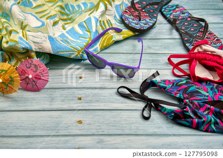 Swimwear, sunglasses, flip flops and beach towel with a tropical print on a wooden table. Swimwear, sunglasses, flip flops and beach towel with a tropical print on a wooden table. 127795098