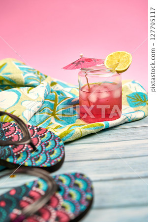 Refreshing pink drink with an umbrella and a slice of lemon on a beach towel with a tropical pattern and flip flops in the foreground with a pink background. Refreshing pink drink with an umbrella and a slice of lemon on a beach towel with a tropical pattern and flip flops in the foreground with a pink background. 127795117