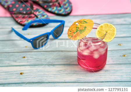 Watermelon and lemon drink with an umbrella and a slice of lemon, blue sunglasses and flip flops on a wooden table with a pink background. Watermelon and lemon drink with an umbrella and a slice of lemon, blue sunglasses and flip flops on a wooden table with a pink background. 127795131