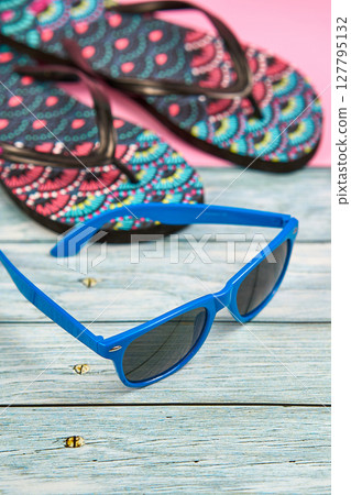 Blue sunglasses and flip flops with a tropical pattern on a wooden table and a pink background. Blue sunglasses and flip flops with a tropical pattern on a wooden table and a pink background. 127795132