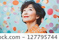 Radiant mature Asian woman with serene expression looks upwards surrounded by colorful falling confetti. Ideal for themes of joy, celebration and contentment 127795744