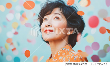 Radiant mature Asian woman with serene expression looks upwards surrounded by colorful falling confetti. Ideal for themes of joy, celebration and contentment Radiant mature Asian woman with serene expression looks upwards surrounded by colorful falling confetti. Ideal for themes of joy, celebration and contentment 127795744