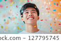 Smiling young Asian man looks up with genuine joy as colorful confetti falls around him, perfect for themes of celebration, youth and achievement 127795745