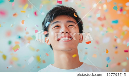 Smiling young Asian man looks up with genuine joy as colorful confetti falls around him, perfect for themes of celebration, youth and achievement Smiling young Asian man looks up with genuine joy as colorful confetti falls around him, perfect for themes of celebration, youth and achievement 127795745