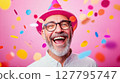 Laughing senior man in a pink party hat and glasses is surrounded by colorful confetti against pink background. Ideal for themes of joy, celebration and milestones 127795747