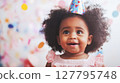Sweet African American girl with curly hair wearing patterned party hat, looks up with happy expression against soft colorful background. Concept: toddler birthday, precious moment 127795748