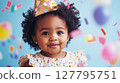 Charming portrait of smiling African-American baby girl wearing festive party hat against blue background with falling colorful confetti and blurred balloons. Joyful birthday celebration 127795751