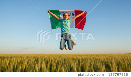 Joyful Woman Leaping With Italian Flag Across Golden Wheat Field Under Clear Blue Sky 127797516
