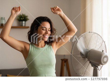 Joyful Woman Celebrates Cool Relief Standing Near Electric Fan in Bright Modern Home Interior 127797898