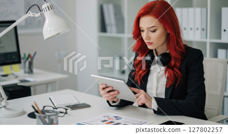 Focused Young Businesswoman with Red Hair Using Tablet in Modern Office Workspace 127802257