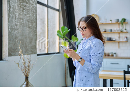 Relaxed mature woman with smartphone cup of drink standing at home 127802825