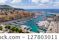 Drone view of the Port of Nice, France, showing luxury yachts, colorful historic buildings, blue Mediterranean Sea, and the coastline under a bright summer sky. 127803011