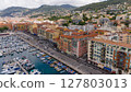 Drone view of colorful buildings along the Port of Nice, France, with yachts, fishing boats, and surrounding hills under cloudy sky. 127803013