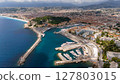 Wide aerial view of the Port of Nice, France, showing the coastline, marina with yachts, city buildings, and surrounding hills on a partly cloudy day. 127803015