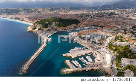 Wide aerial view of the Port of Nice, France, showing the coastline, marina with yachts, city buildings, and surrounding hills on a partly cloudy day. 127803015