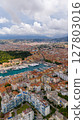 Vertical aerial view of the Port of Nice, France, showing moored yachts, colorful buildings, and surrounding hills under a partly cloudy sky. 127803016