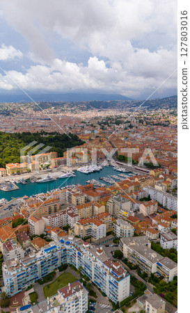 Vertical aerial view of the Port of Nice, France, showing moored yachts, colorful buildings, and surrounding hills under a partly cloudy sky. 127803016