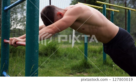 Young muscular athlete doing exercise on triceps at sport equipment outdoor. Male shirtless athlete working out at sport ground. Strong sporty man training at nature. Concept of healthy lifestyle Young muscular athlete doing exercise on triceps at sport equipment outdoor. Male shirtless athlete working out at sport ground. Strong sporty man training at nature. Concept of healthy lifestyle 127803019