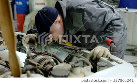 Experienced auto mechanic wearing gloves and uniform carefully inspecting car engine compartment in a professional garage workshop, performing diagnostics and maintenance 127803241