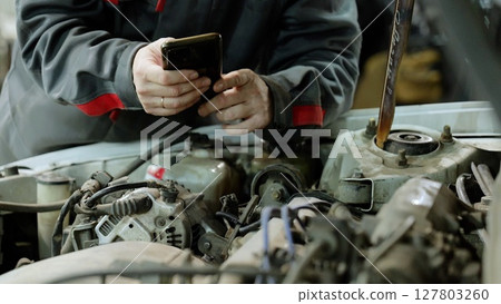 Car mechanic checking vehicle engine using smartphone, displaying digital diagnostic technology in automotive maintenance 127803260