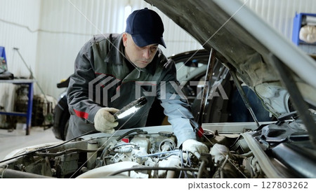 Professional mechanic inspecting vehicle engine, wearing protective gear and using flashlight while performing detailed diagnostic work inside modern automotive repair facility Professional mechanic inspecting vehicle engine, wearing protective gear and using flashlight while performing detailed diagnostic work inside modern automotive repair facility 127803262