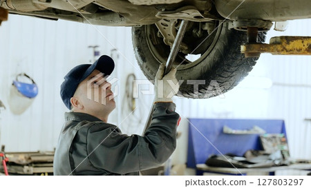 Skilled mechanic fixing vehicle suspension with wrench, working intently under raised car in professional automotive repair workspace 127803297