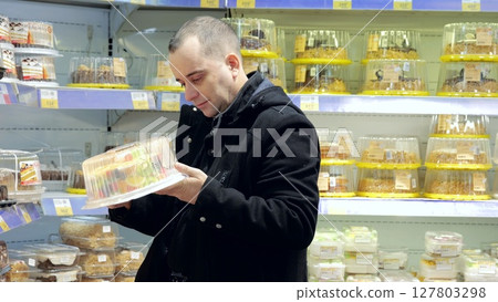 Male shopper browsing packaged fruit cakes, comparing selections within refrigerated bakery display, contemplating sweet dessert purchase at grocery store 127803298