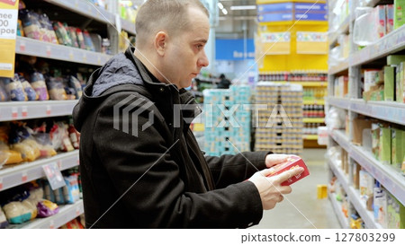 Consumer carefully examining product label on red packaging while grocery shopping, focusing on ingredient details and nutritional information in supermarket aisle Consumer carefully examining product label on red packaging while grocery shopping, focusing on ingredient details and nutritional information in supermarket aisle 127803299