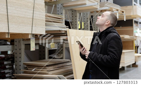 Carpenter checking wooden boards, selecting quality materials using smartphone, standing amid stacked timber and plywood in warehouse setting 127803313