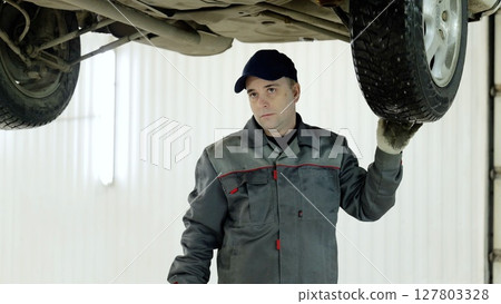 Professional mechanic in uniform checking car wheel while working underneath lifted vehicle, maintaining safety standards in well lit automotive repair workspace Professional mechanic in uniform checking car wheel while working underneath lifted vehicle, maintaining safety standards in well lit automotive repair workspace 127803328