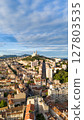 Drone aerial view of the city of Marseille, France, with the iconic Notre-Dame de la Garde basilica on a hilltop surrounded by urban buildings under a partly cloudy sky. 127803535