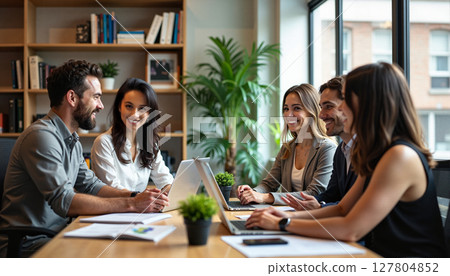 A group of professionals gathered around a table with laptops and papers, engaged in a collaborative discussion in an office setting. 127804852