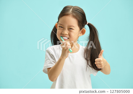 Cute Chinese little girl having perfect smile brushing her teeth with plastic toothbrush on blue background, keep thumb raised, studio portrait. 127805000