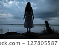 Mysterious woman in white dress standing by a dark lake under stormy sky 127805587