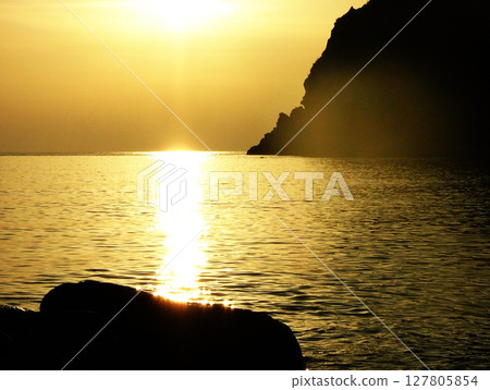Stunning golden sunset over the sea in Calabria, Italy, with soft sunlight reflecting on the water and the curved silhouette of mountain cliffs in the distance. Serene coastal landscape with warm 127805854