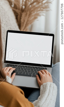 Woman sitting on a comfortable sofa at home, typing on a laptop computer with a blank white screen, perfect for showcasing designs and mockups in a cozy setting Woman sitting on a comfortable sofa at home, typing on a laptop computer with a blank white screen, perfect for showcasing designs and mockups in a cozy setting 127807706