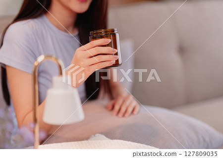 Young Asian Woman Enjoying Relaxing Scented Candle Ambiance with Modern Warmer Lamp for Home Wellness and Stress Relief Young Asian Woman Enjoying Relaxing Scented Candle Ambiance with Modern Warmer Lamp for Home Wellness and Stress Relief 127809035