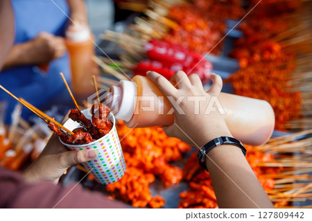 Filipino street food skewers with red sauce in paper cup, hand pouring sauce at outdoor market 127809442