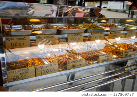 Traditional Filipino cooked meals displayed behind glass at carinderia food stall with handwritten price signs 127809757
